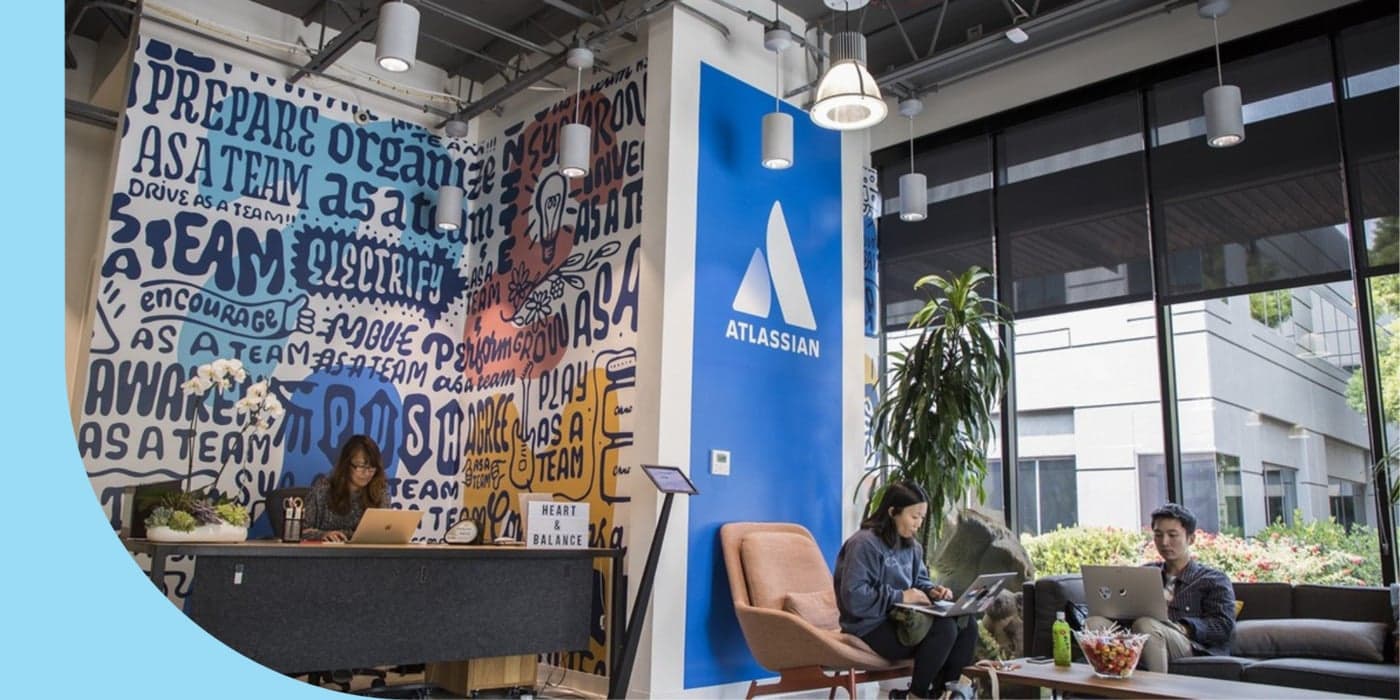 Three people sitting in a brightly lit office lounge, working on their laptops. The person on the far left wears a dark floral print blouse. The person in the middle wears a blue sweatshirt and black leggings. The person on the far right wears a plaid shirt and khaki pants.