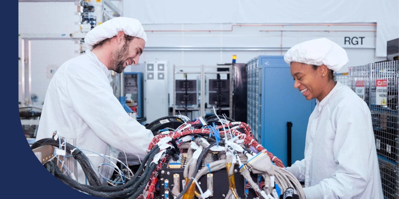 Two people facing each other, wearing protective white jumpsuits and white bonnets. The person on the left has facial hair. The person on the right wears earrings. They are working on a piece of machinery with many wires and cables.