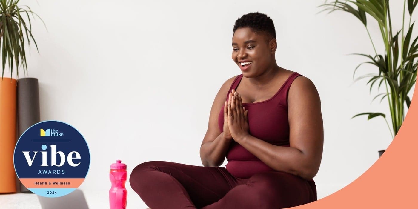 A woman in athletic clothes smiles as she sits cross legged with her palms together in front of her chest in a yoga studio